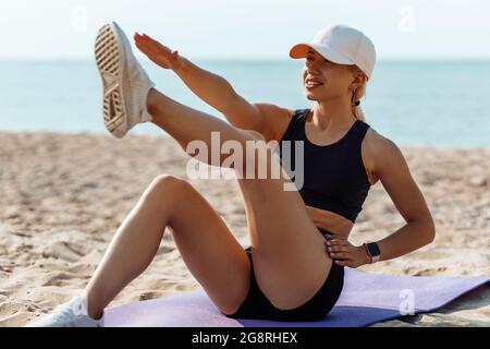 Young sportive woman doing sports outdoors on the beach, lying on a yoga mat, doing abs exercises, Young attractive woman doing abs workout by the sea Stock Photo