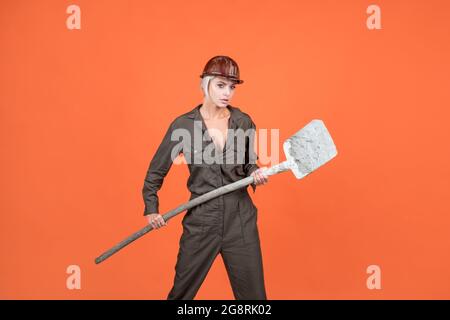 woman hard worker in protective helmet and boilersuit hold shovel on orange background, labour day Stock Photo