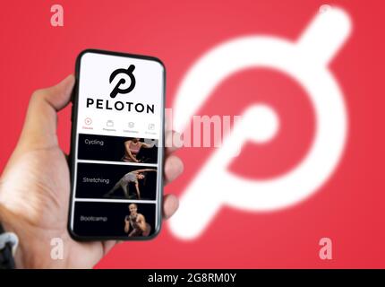 New York, USA, July 2021: A hand holding a phone with the Peloton app on the screen. Red background with blurred Peloton logo. Peloton is an American Stock Photo