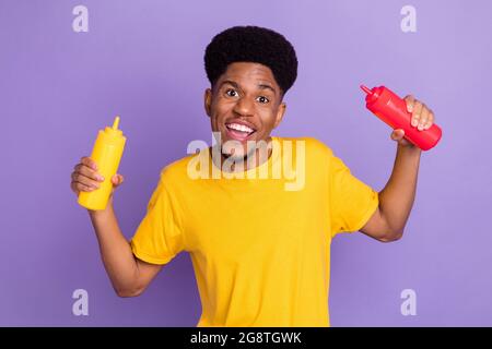 Portrait of attractive carefree cheerful guy holding ketchup bottles having fun isolated over violet purple color background Stock Photo