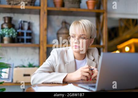 Photo of a pensive woman in front of a laptop monitor. She works with documents and papers at home - checks the reports of tax forms online during qua Stock Photo