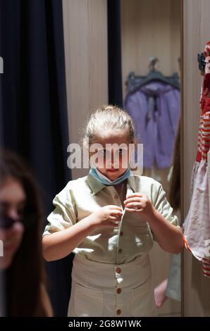 Little girl in a protective medical mask chooses school uniform store.  Girl trying on clothes in a fitting room. Preparing for school. Prevention of Stock Photo