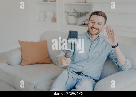 Young man enjoying video chat with family on cellphone while sitting on sofa in living room