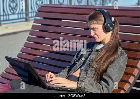 A woman with a laptop sits on an outdoor bench and works online Stock Photo