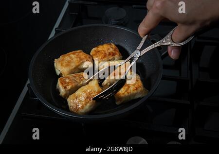 Hand of woman with kitchen tongs is turning stuffed cabbage rolls on frying pan on black kitchen stove . Freshly fried rolls are covered by delicate c Stock Photo