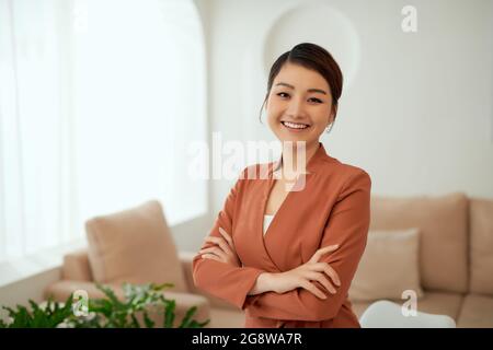 Asian working woman who wears black suit and cross arms stands in white office room, Stock Photo