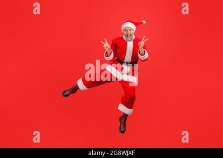 Full length portrait of happy elderly man with gray beard wearing santa claus costume jumping showing v gesture with fingers, celebrating winter holid Stock Photo