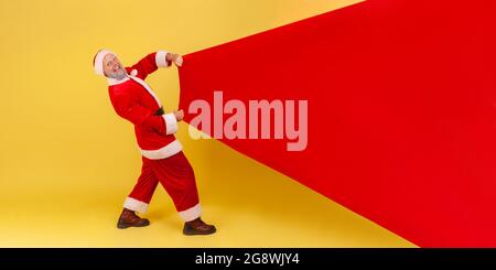 Extremely happy smiling elderly man with gray beard wearing santa claus costume pulling huge bag of gifts, copy space for advertisement. Indoor studio Stock Photo