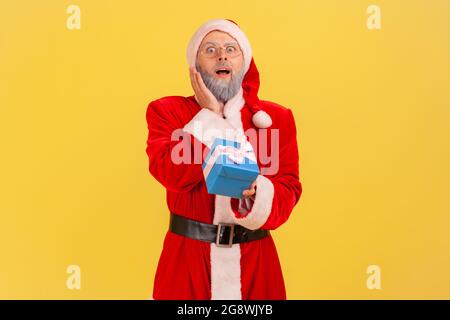 Extremely surprised elderly man with gray beard wearing santa claus costume holding wrapped blue present box, has gift on new year eve. Indoor studio Stock Photo