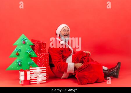 Surprised elderly man wearing santa claus costume sitting on floor under decorated christmas tree with gift boxes and big red bag with holidays presen Stock Photo