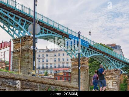 A 19th Century footbridge of iron resting on stone columns with braced iron arches.  Seagulls are nesting on the columns and a white building is beyon Stock Photo
