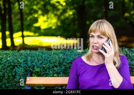 Defocus angry caucasian blond woman talking, speaking on the phone outside, outdoor. 40s years old woman in purple blouse in park on bench. Adult Stock Photo