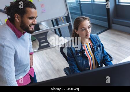 Two diverse creative colleagues sitting at desk using computer and smiling Stock Photo