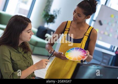 Two diverse female creative colleagues sitting at desk talking Stock Photo