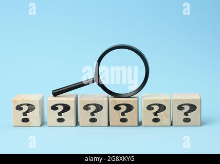 wooden cubes with question marks and a magnifying glass on a blue background. The concept of finding answers to unknown questions, solving a problem. Stock Photo