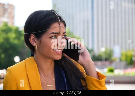 Close up of a young indian businesswoman wearing yellow suit jacket talking on the smartphone sitting outdoor. Stock Photo