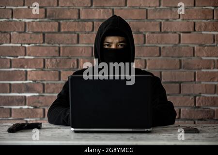 Cyber robber in black hood with notebook computer, gun and mobile phone stare at camera. Concept for internet crimes and illegal manner. Stock Photo