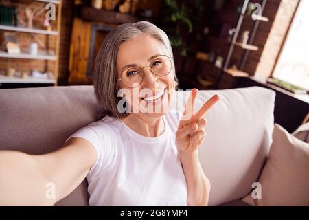 Self-portrait of attractive cheerful grey-haired woman sitting on sofa showing v-sign having fun at home house indoors modern brick interior Stock Photo