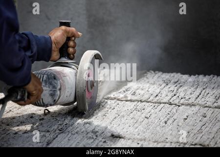 Hands of worker catching and using electric cutting machine tool to cut concrete floor with dirty dust spreading in air, copy space on the right. Stock Photo