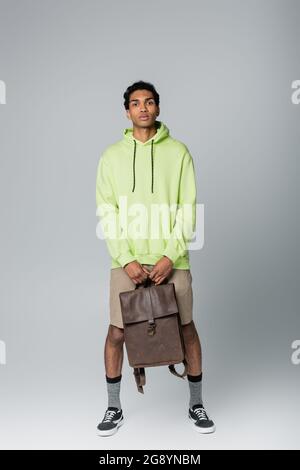 full length view of african american guy in green hoodie standing with leather backpack isolated on grey Stock Photo