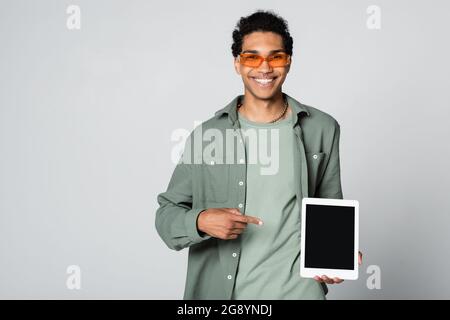 cheerful african american guy in orange eyeglasses pointing at digital tablet isolated on grey Stock Photo