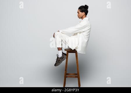 side view of african american man sitting on high stool in white suit and sneakers on grey background Stock Photo