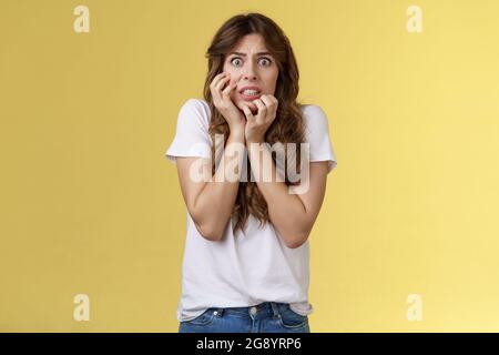 Worried insecure nervous european girl biting fingernails stare camera anxious panic touch cheek timid intense stooping stand yellow background scared Stock Photo