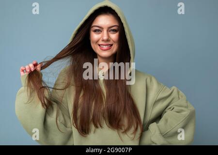 Portrait of attractive young caucasian smiling brunette woman model in trendy khaki hoodie and wearing hood isolated on blue background with copy Stock Photo