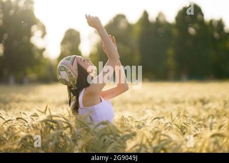 Young woman wearing bandana standing with arms raised in wheat field during sunny day Stock Photo