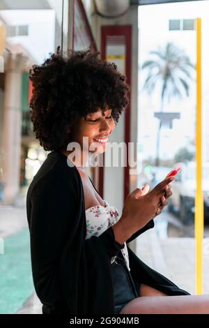 Smiling Afro woman using smart phone at bus top Stock Photo