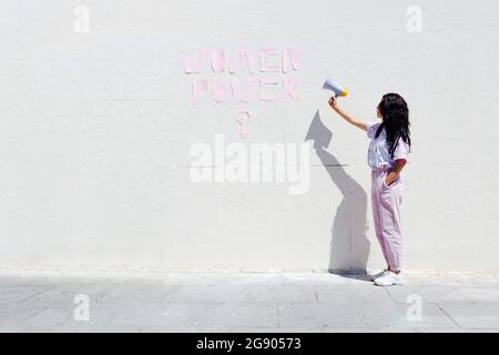 Young woman with hand in pocket standing by text on wall while holding megaphone during sunny day Stock Photo