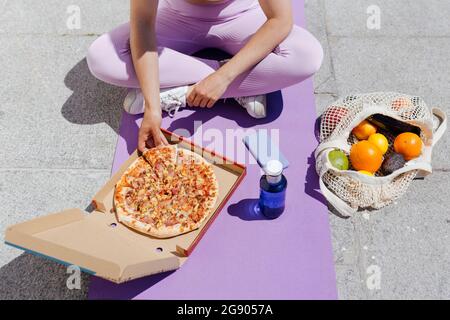 Sportswoman having pizza while sitting cross-legged on exercise mat Stock Photo