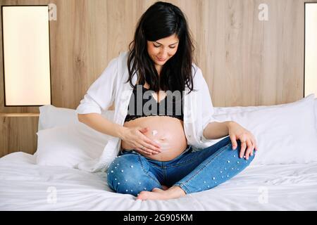 Pregnant woman applying moisturizer on belly while sitting on bed in bedroom Stock Photo