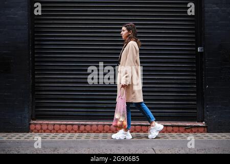 Young woman holding mesh bag while walking by closed shutter Stock Photo