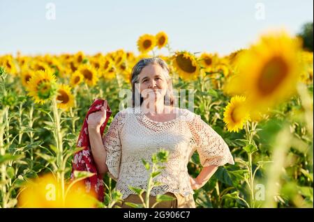 Senior woman standing with hand on hip in sunflower field Stock Photo
