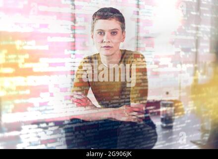 Businesswoman sitting on desk seen through computer codes on glass in office Stock Photo