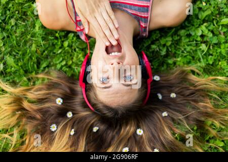 Shocked young woman listening music through headphones with flowers in hair lying on grass Stock Photo