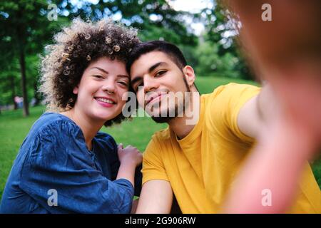 Smiling curly haired woman sitting with boyfriend at park Stock Photo