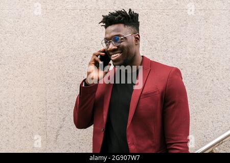 Smiling young businessman talking on mobile phone while standing in front of wall Stock Photo