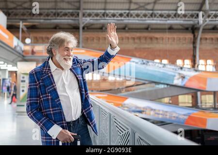 Mature man waving hand while standing by railing at station Stock Photo
