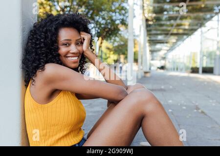 Smiling young woman with curly hair sitting by pillar Stock Photo