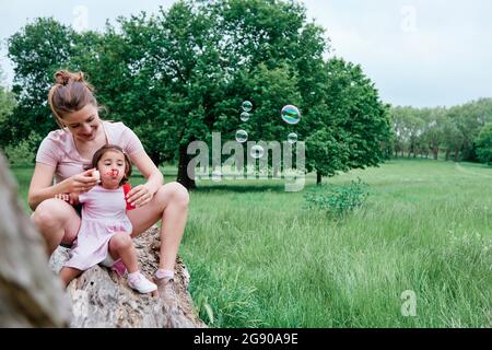 Daughter blowing bubbles with mother sitting on log in park Stock Photo
