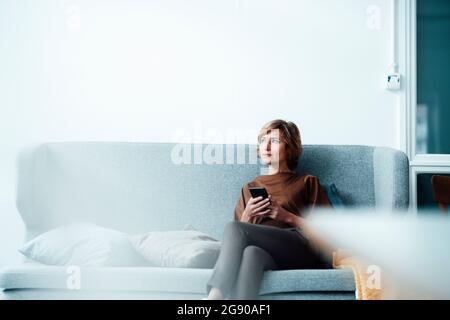 Thoughtful businesswoman with mobile phone sitting on sofa Stock Photo