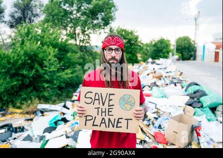 Serious man holding save the planet cardboard on garbage dump Stock Photo