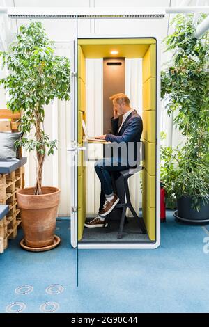 Male professional using laptop while talking on mobile phone in soundproof booth Stock Photo