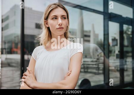 Serious woman with arms crossed looking away in front of glass wall Stock Photo