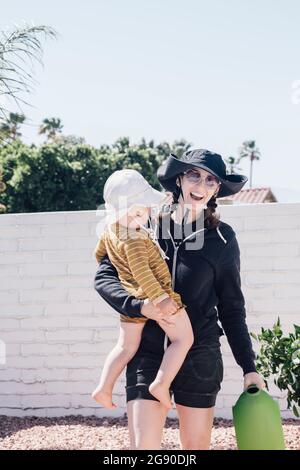 Happy woman with watering can carrying son during sunny day in patio Stock Photo