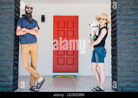 Smiling woman carrying dog while man with arms crossed standing in front of house Stock Photo