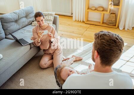 Smiling young woman photographing through mobile phone while man carrying daughter at home Stock Photo