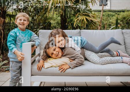 Boy standing by sofa while sisters enjoying at backyard Stock Photo
